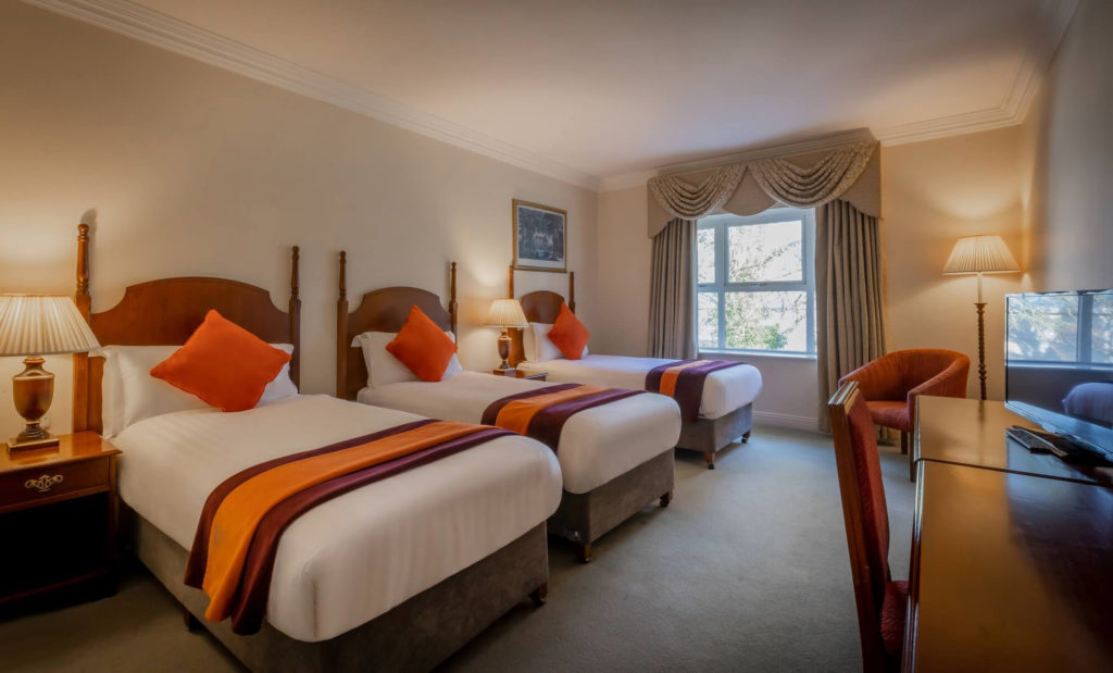 Hotel room with two single beds, white linens, orange pillows and throws, bedside lamps, a window with curtains, an armchair, and a desk with a TV.