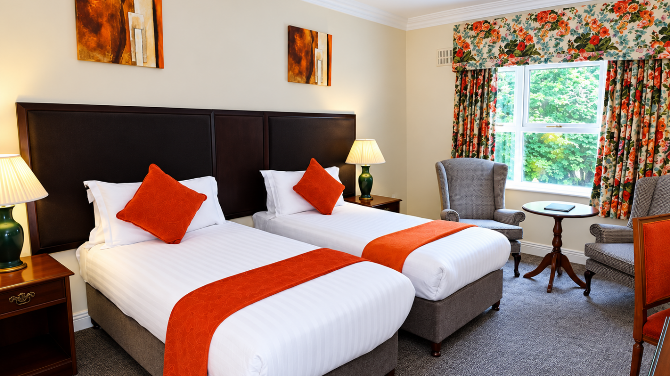 Bright twin-room with crisp white linens, orange throws and cushions, armchairs by a floral-curtained window, bedside lamps and wood furnishings