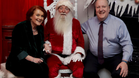 Three adults pose indoors; one dressed as Santa Claus sits on a white throne between a woman in green and a man in a blue shirt. Holiday decorations are visible in the background.