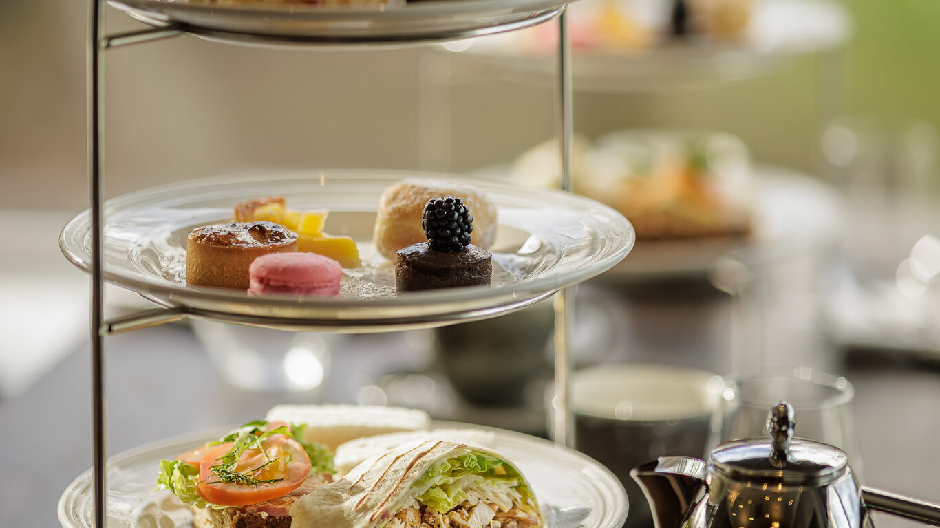 Elegant afternoon tea with three-tiered stand featuring sandwiches, scones, and desserts, with a scenic garden view.