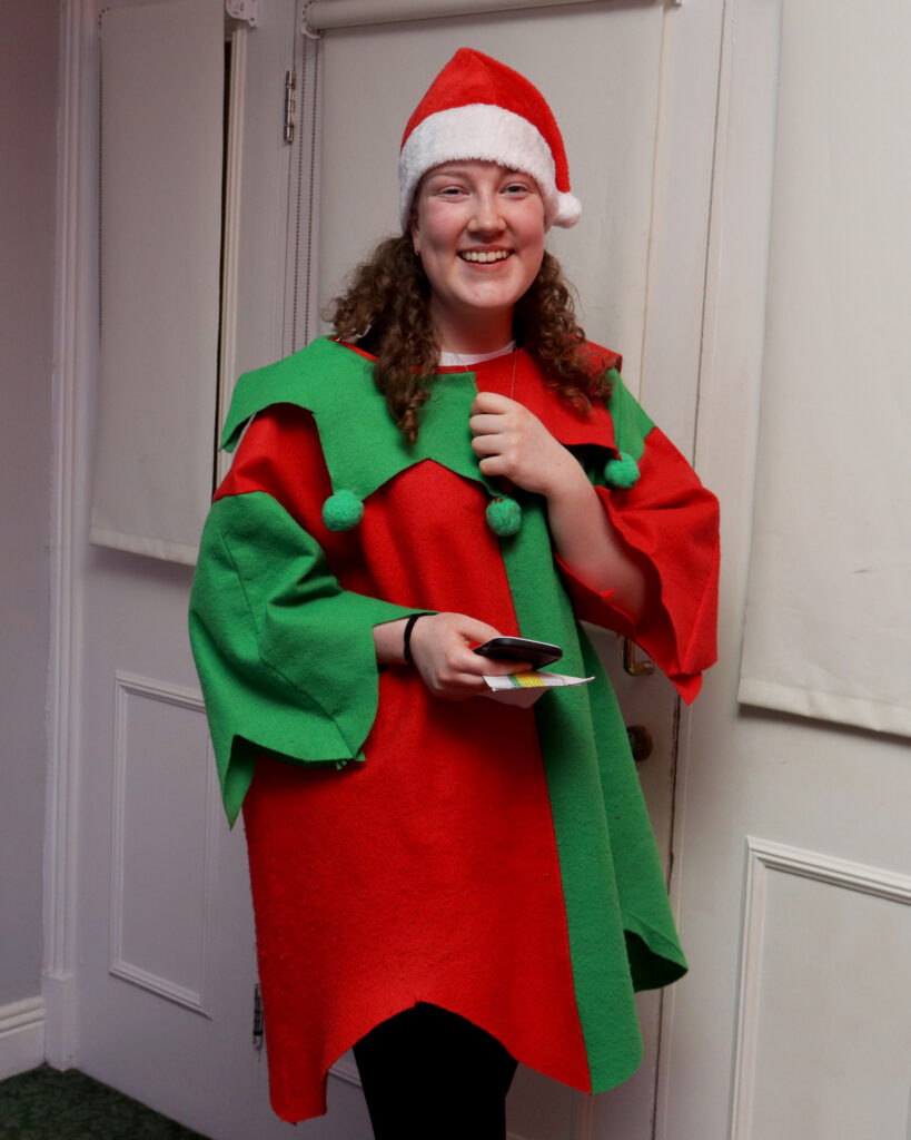 IMG_0979 edit A person wearing a Santa hat and a red and green festive costume stands indoors, smiling and holding a plate of food.