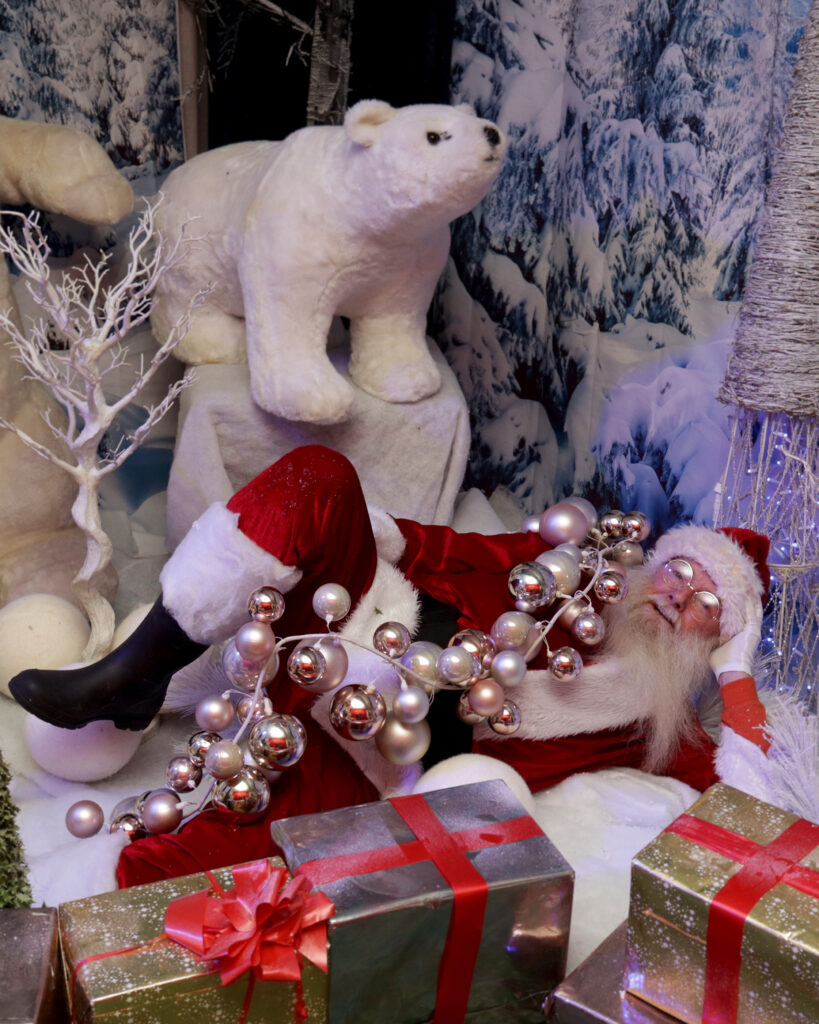 IMG_0980 edit Santa Claus lies on artificial snow among wrapped gifts, tangled in silver baubles, with a large polar bear decoration and wintry forest background.