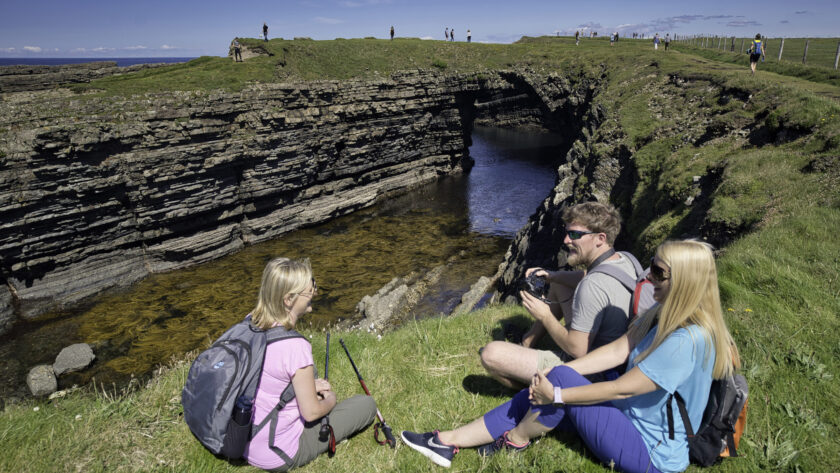Guests enjoying a scenic coastal hike, sitting on lush green grass with stunning cliff views under a clear blue sky.