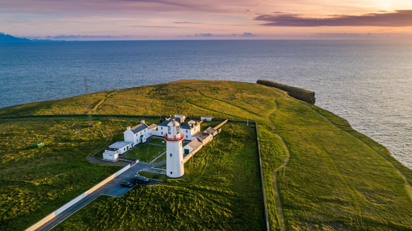 Stunning lighthouse hotel on clifftop with ocean views, green pastures, and sunset skies offering a serene coastal retreat.