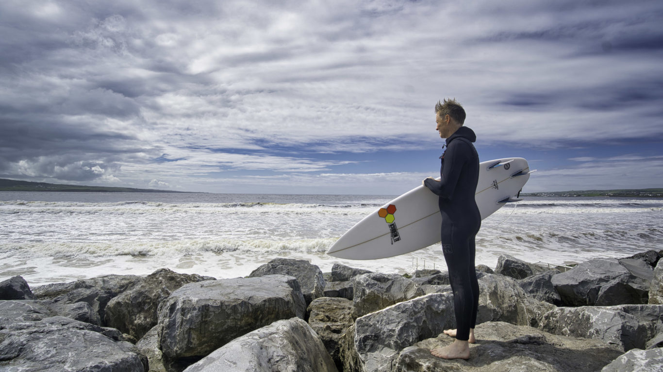 Surfer in wetsuit stands on rocky shore, holding a surfboard, gazing at the scenic ocean waves under a cloudy sky.