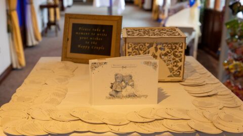 Elegant wedding guest book table with ornate box and cards inviting guests to leave wishes, set in a warm hotel hallway.