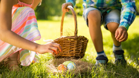 Children enjoying an Easter egg hunt on lush hotel lawns, wearing colourful clothes, with a wicker basket and sunny skies.