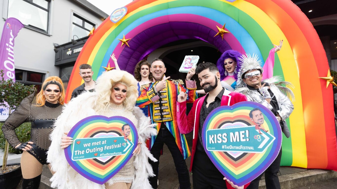A group of people in colorful costumes pose in front of a large inflatable rainbow arch at a festival, holding heart-shaped signs with messages.