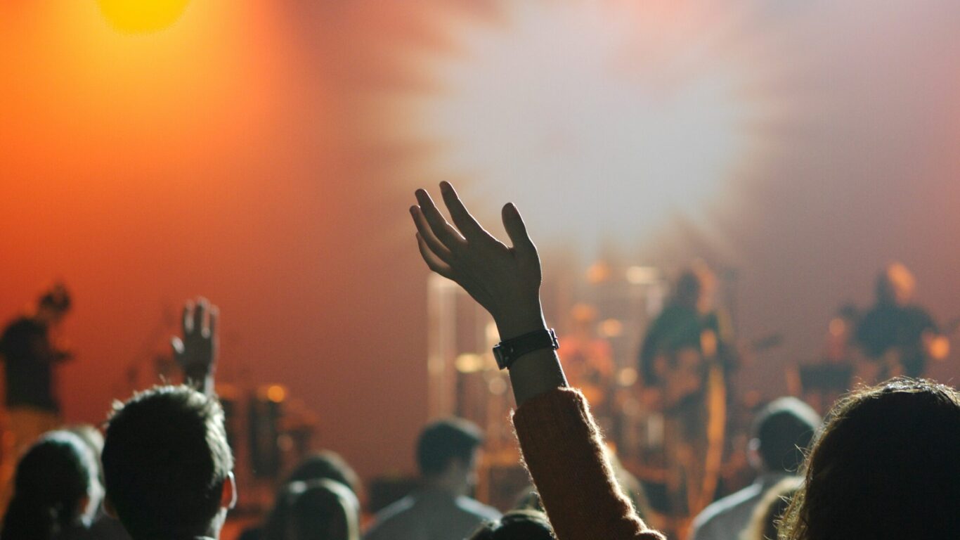 People in an audience raise their hands toward a brightly lit stage where musicians perform, with orange and white lighting effects in the background.