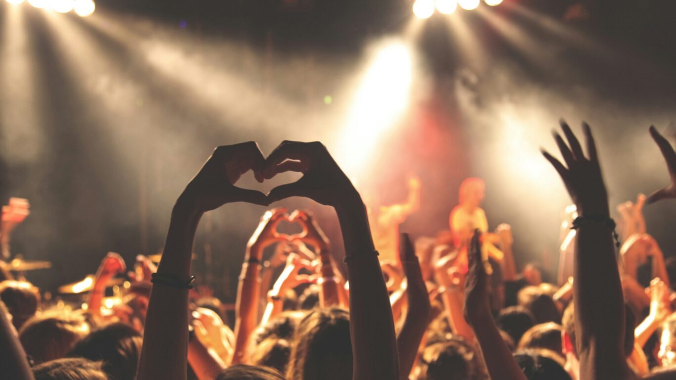 A crowd at a concert raises their hands, with some making heart shapes, while bright stage lights illuminate the scene and performers appear slightly blurred in the background.