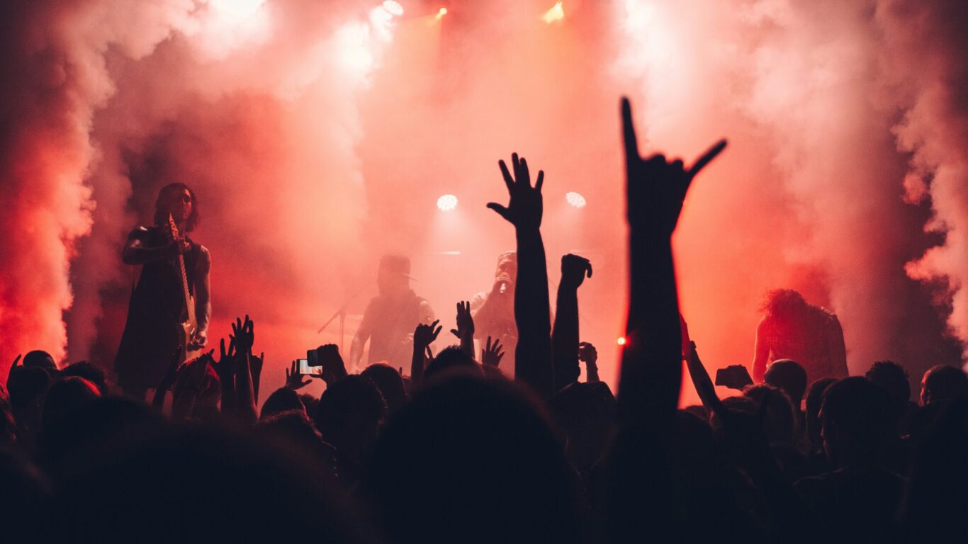 A crowd raises their hands at a rock concert with red stage lights and smoke, while a band performs on stage in the background.