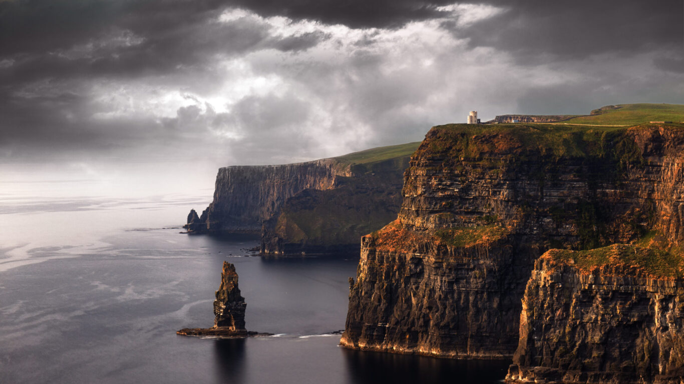 View of the Cliffs of Moher in Ireland, featuring steep rocky cliffs rising above the ocean under a cloudy sky, with a sea stack visible in the water.