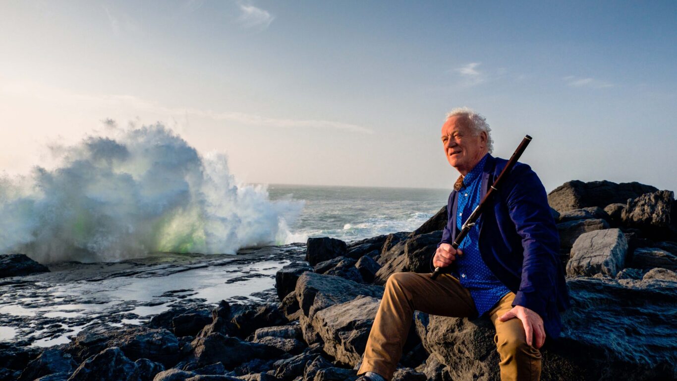 An older man sits on rocky shore holding a flute, with ocean waves crashing dramatically against rocks in the background under a clear sky.