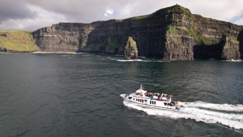 A white tour boat travels near the base of tall, rocky sea cliffs with green tops under a cloudy sky.