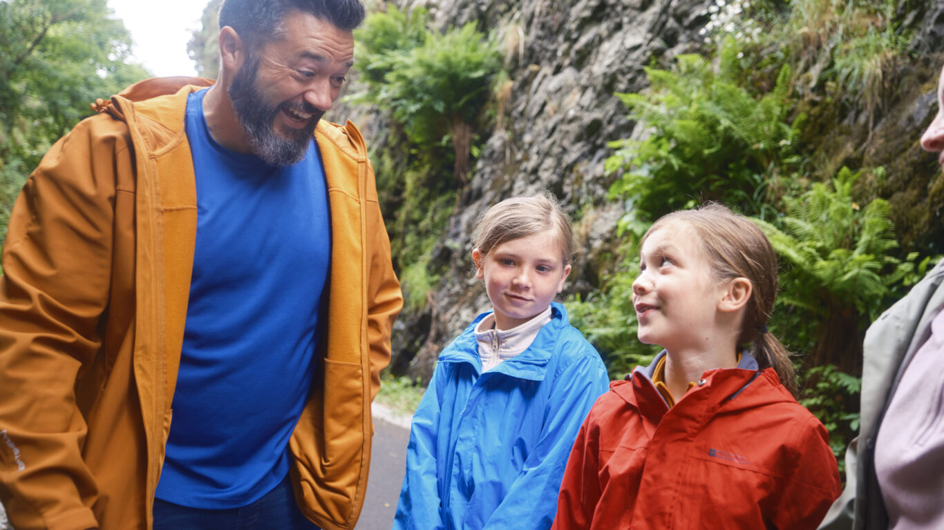 An adult and two children, all wearing jackets, stand on a paved path outdoors with trees and rocky cliffs in the background.