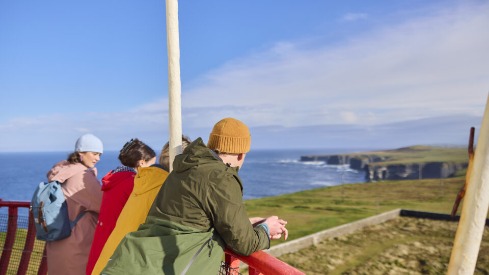 Four people in outdoor jackets stand on a red railing platform looking out over coastal cliffs and the ocean under a blue sky.