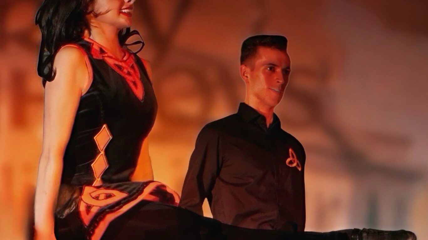Two dancers perform an Irish dance, both mid-air with arms by their sides, wearing traditional costumes against a blurred stage background.