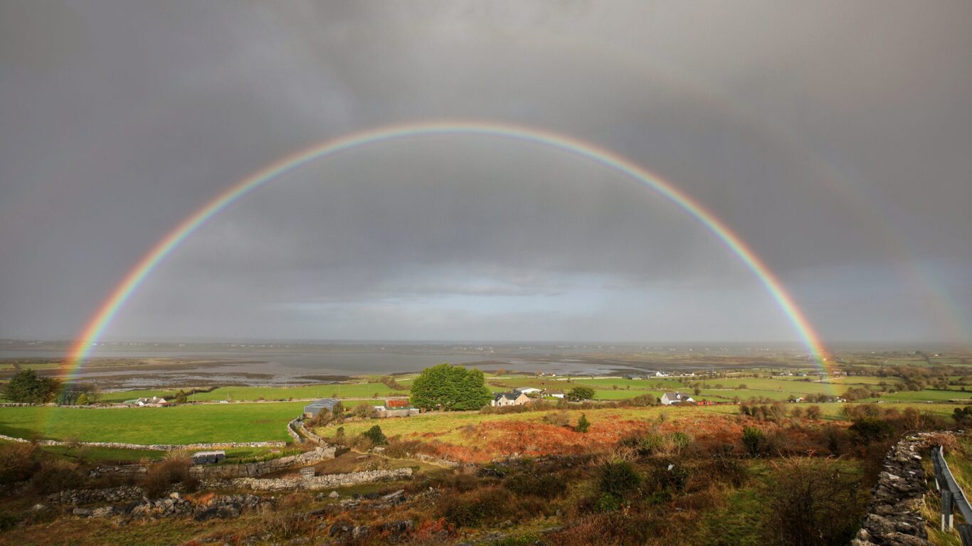 A vivid rainbow arcs over a rural landscape with fields, scattered houses, and cloudy skies in the background.