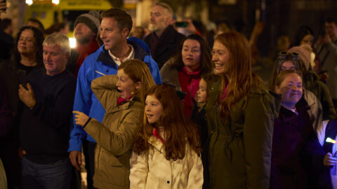A group of people, including adults and children, stand outdoors at night, looking in the same direction with expressions of surprise and excitement.