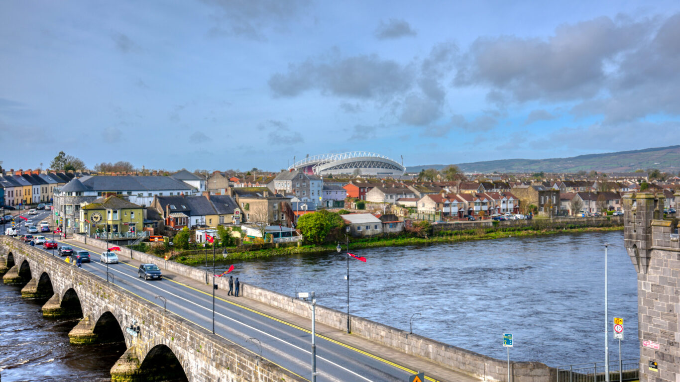 Stone bridge over a river with cars and pedestrians, residential houses and a stadium in the background, under a partly cloudy sky.
