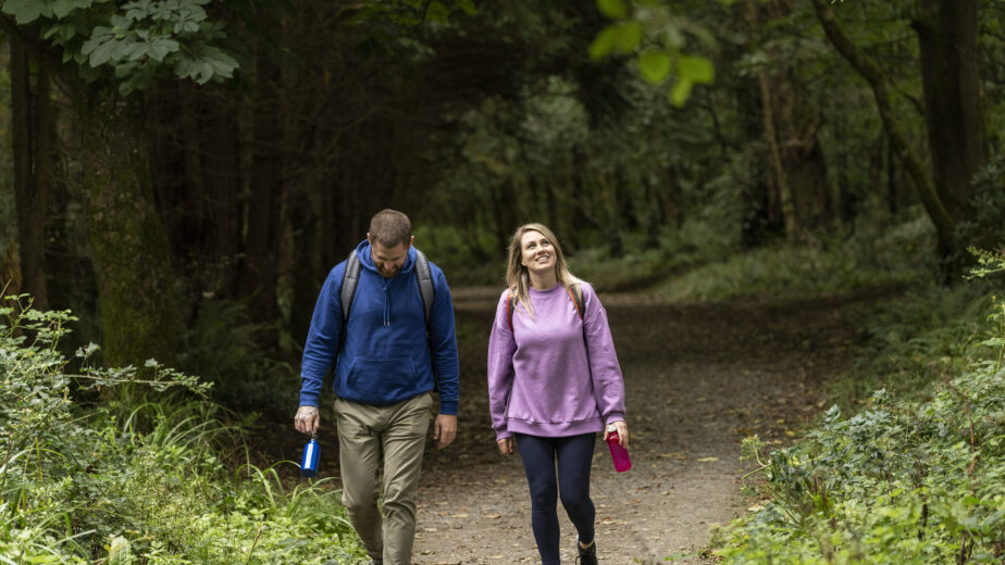 Two people with backpacks walk along a forest path; one wears a blue hoodie and holds a blue bottle, the other wears a purple sweatshirt and holds a pink bottle.