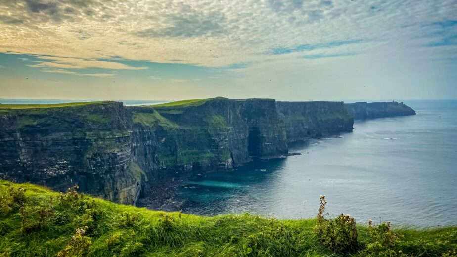 Coastal cliffs with green grass above, overlooking the ocean under a partly cloudy sky.