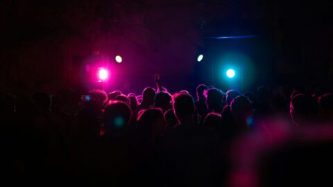 A crowd of people stands in a dark space, lit by bright pink and blue stage lights, with some raised hands visible in the foreground.