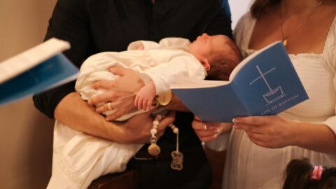 An adult holds a baby dressed in white while another adult holds a blue booklet titled 