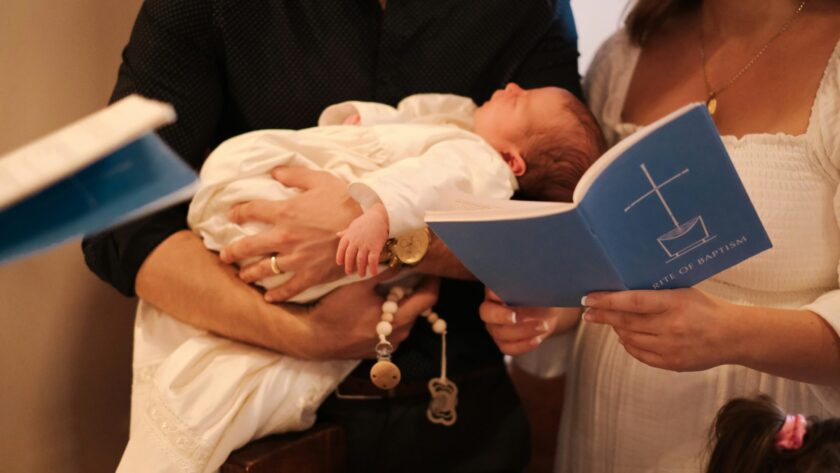 An adult holds a baby dressed in white while another adult holds a blue booklet titled