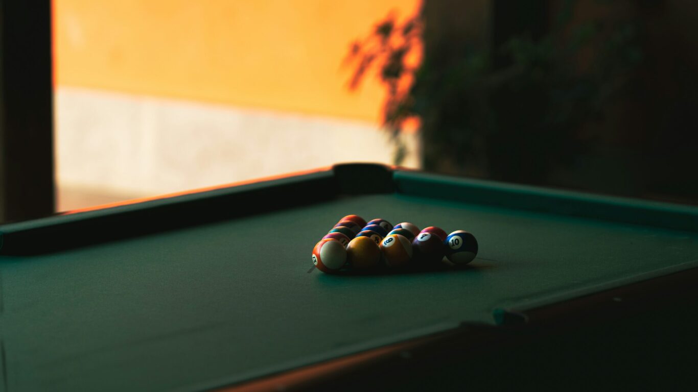 A racked set of billiard balls sits on a green pool table in a dimly lit room with an orange wall in the background.