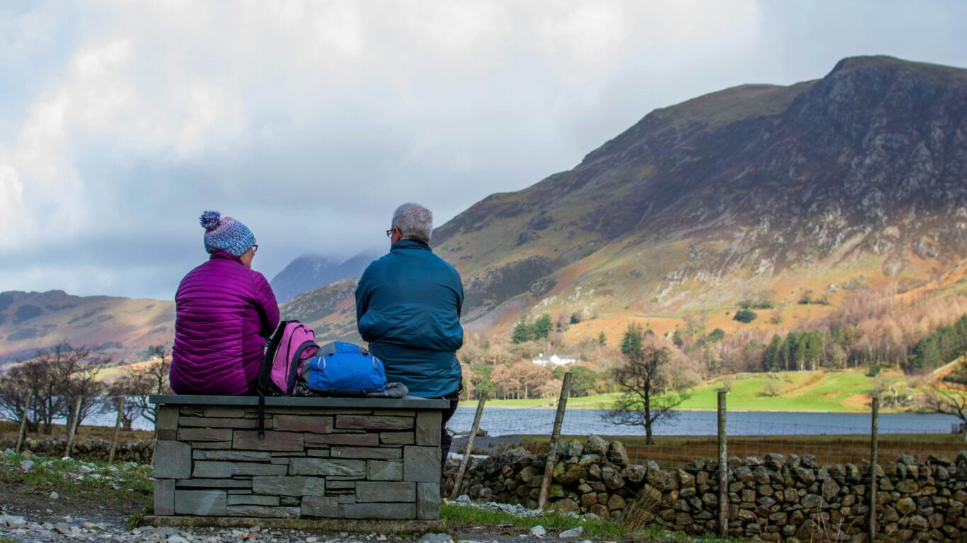 Two people sit on a stone bench facing a scenic landscape with hills, a lake, and cloudy sky. They wear jackets and backpacks.