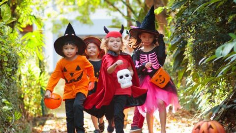 Five children in Halloween costumes, including witches, a pumpkin, and a devil, walk outside on a path holding treat buckets.