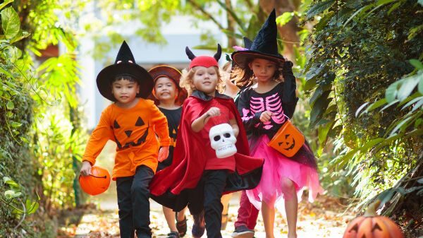 Five children in Halloween costumes, including witches, a pumpkin, and a devil, walk outside on a path holding treat buckets.