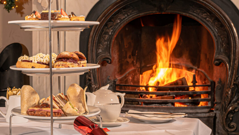 Three-tiered tray with assorted pastries and sandwiches on a table set for tea, in front of a lit fireplace and decorated Christmas tree.