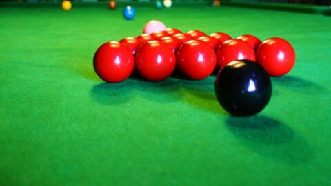 A snooker table with red, black, and colored balls arranged, viewed from one end under a ceiling light in a dimly lit room.