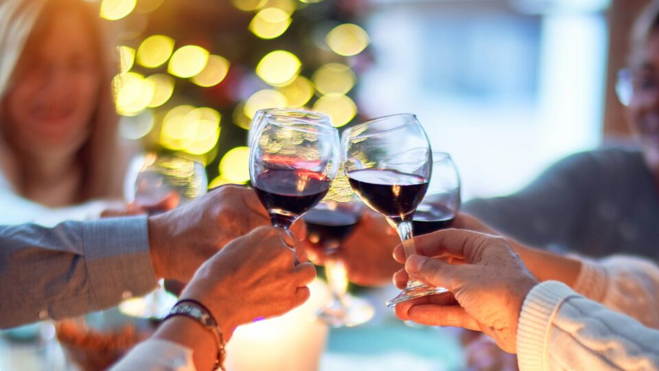 People sit around a festive table with food, clinking wine glasses in a toast. Lit candles and a decorated Christmas tree are visible in the background.