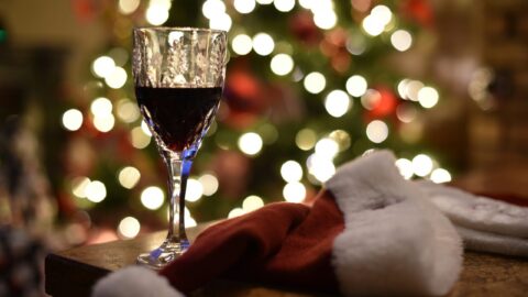 A glass of red wine sits on a table next to a Santa hat, with a decorated Christmas tree and blurred lights in the background.