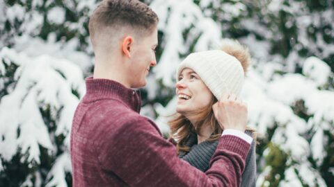 A man and woman dressed in winter clothing stand close together outdoors, smiling at each other with snow-covered trees in the background.