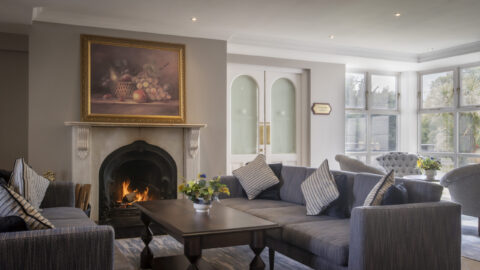 Cosy hotel lounge with plush sofas, roaring fireplace, and large windows offering garden views; perfect for relaxation.