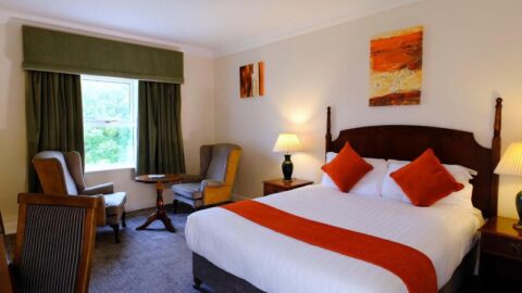 Spacious hotel room with elegant decor, a king-size bed, plush armchairs, large window, and warm lighting.