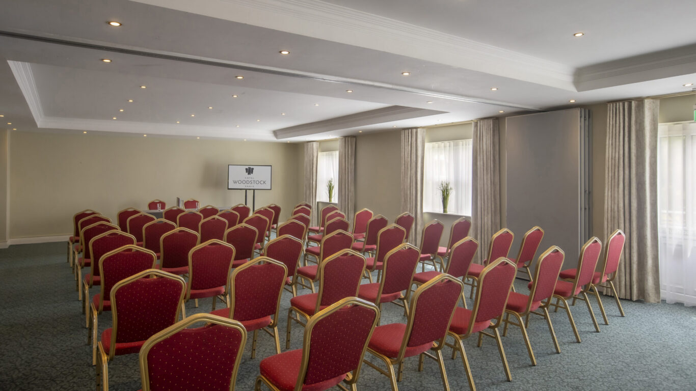 Light-filled conference room with rows of red cushioned chairs, projector screen, natural light, curtains and carpeted floor &mdash; ideal for meetings.