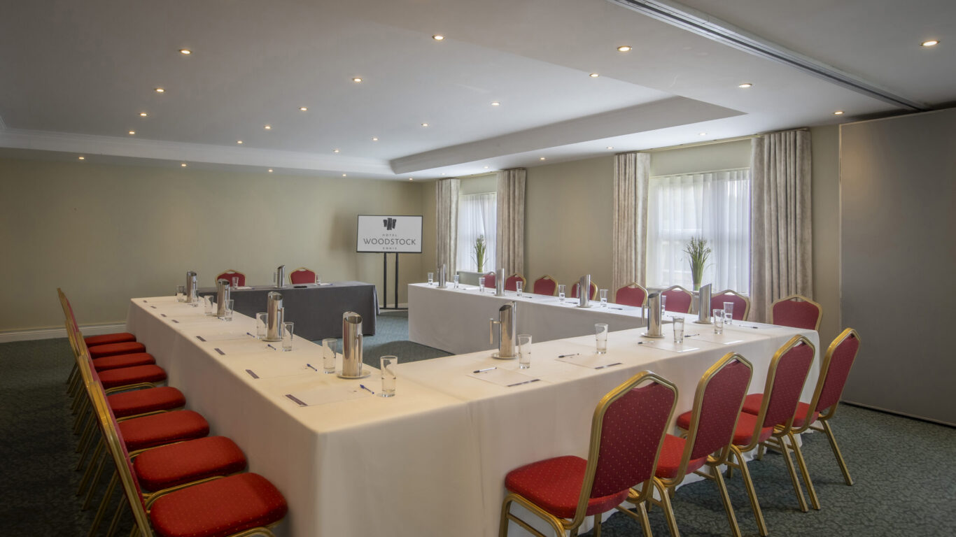U-shaped conference room with white-clothed tables, red banquet chairs, water jugs, notepads and screen, bright natural light and carpeting
